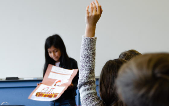 Image of a classroom with teacher holding up a poster and child putting their hand up