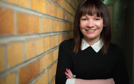 Dr Deborah O'Neil is wearing a black jumper with white collar and standing against a brown brick wall. She had dark brown hair to her shoulders and a fringe.