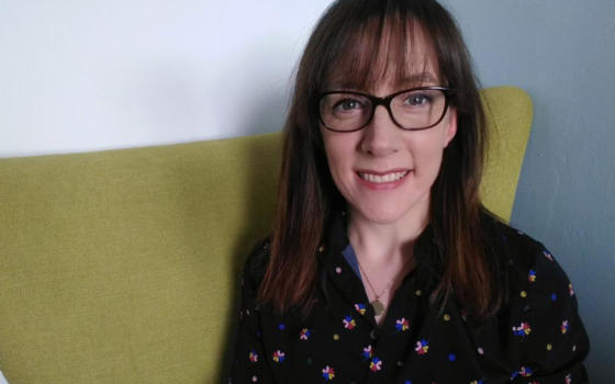 Dr Siobhan O'Connor is wearing a floral blouse and has long dark hair and dark glasses. She is sitting on a yellow sofa