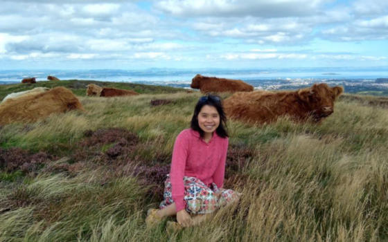 Dr Stella Chan is sitting outside in a field wearing a pink cardigan and skirt. She has dark hair cut into a bob and she is smiling. Highland Cattle are visible in the background