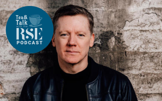 Fergus Linehan is male with short brown hair. He is leaning against a grungy, cracked wall. Wearing all black including a shiny leather jacket.