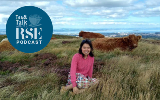 Dr Stella Chan is sat in on a hill in front of a herd of highland cows - she is wearing a pink jumper with a flower knee length skirt