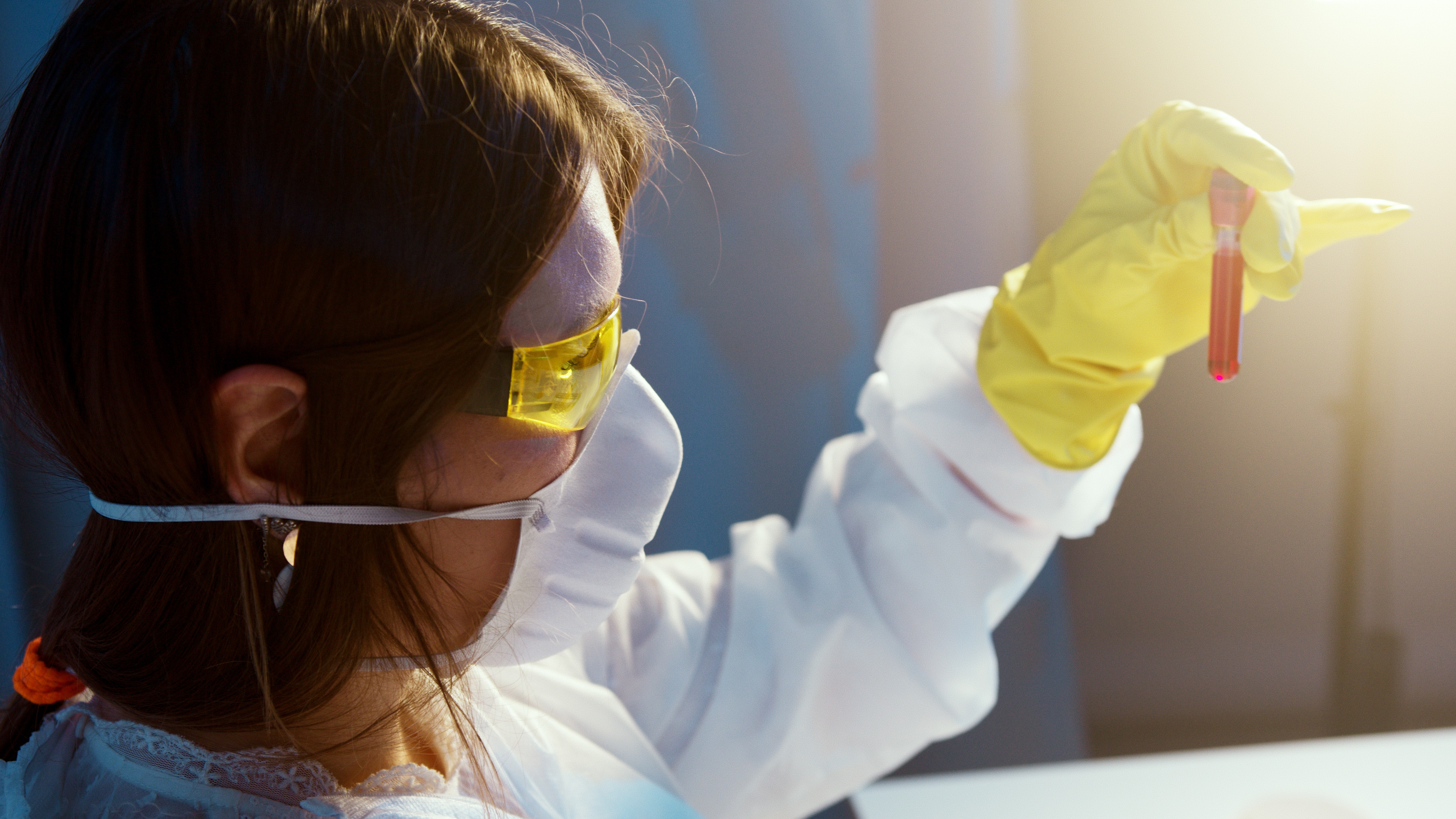 A female scientist wearing PPE including eye protection and a face mask examines a test tube.