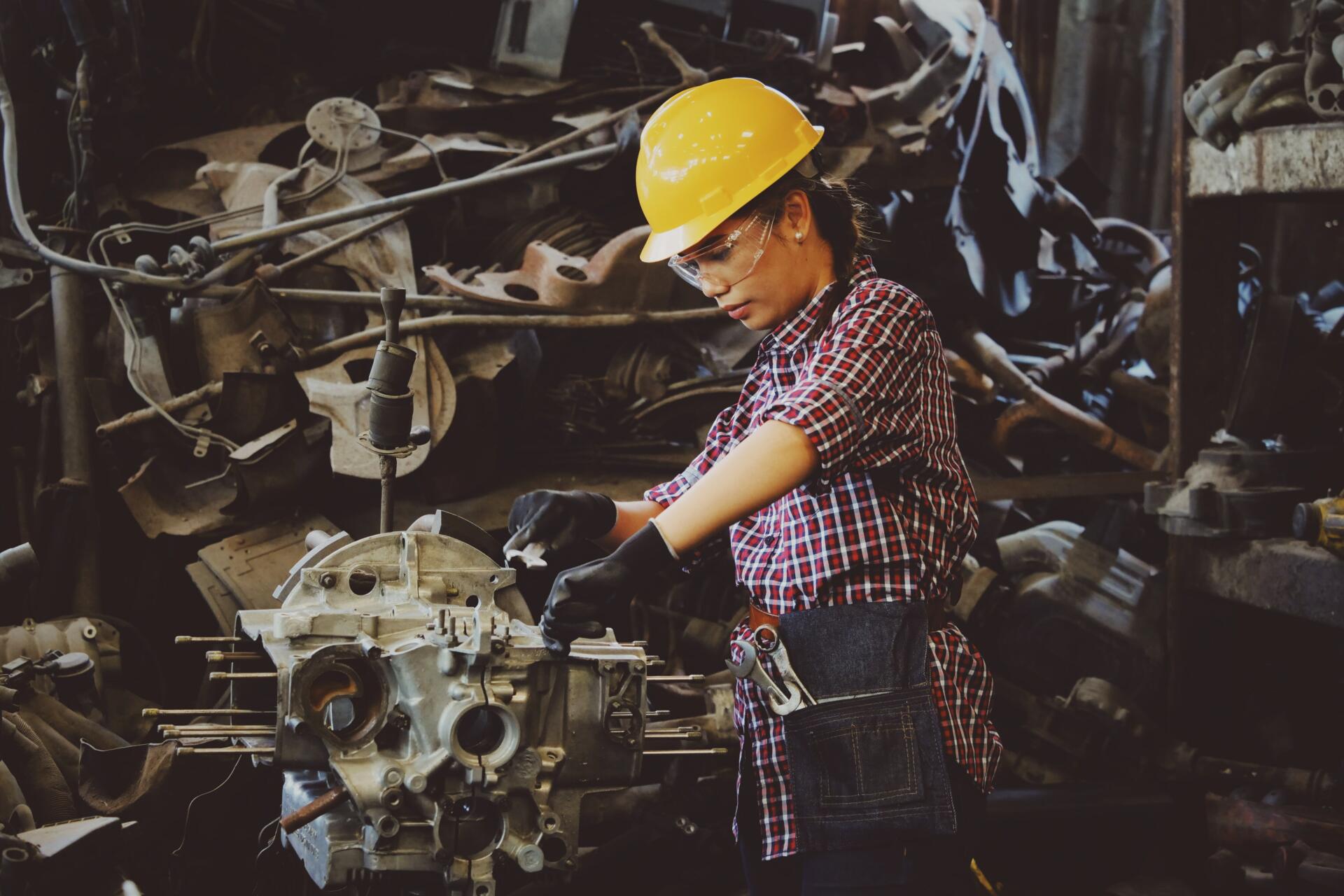 A woman in a checked shirt and a hard hat works on part of a machine.