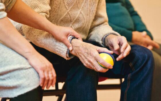 An elderly hand holds a ball, and another hand rests on the arm in comfort.