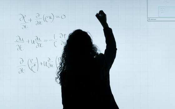 The silhouette of a woman is visible writing equations on a white board.