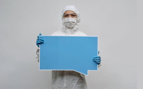 A figure in full PPE including blue gloves and a face mask holds a large blue speech bubble.