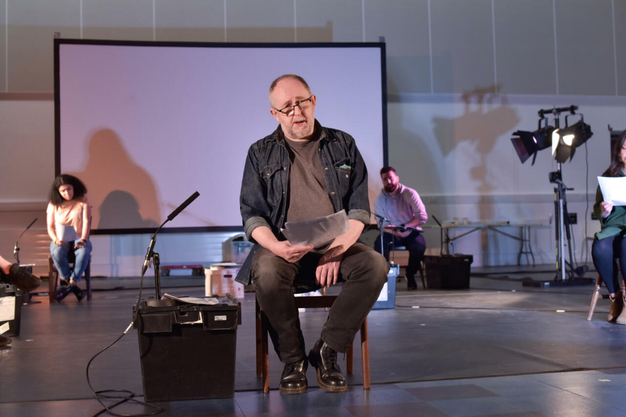 A man sits on a chair on stage and reads from a script.