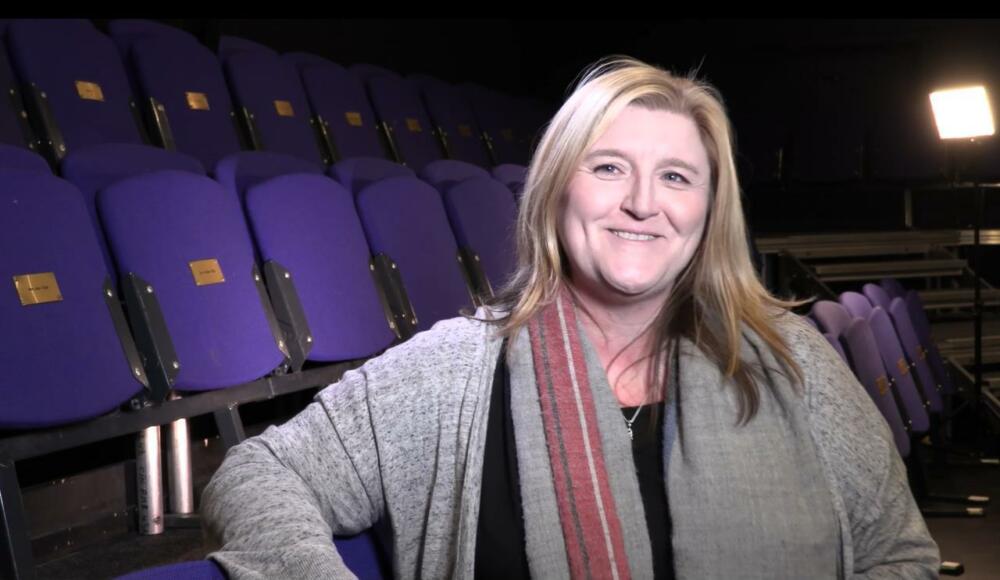 Lead artist Nicola McCartney stands in front of empty chairs smiling at the camera.